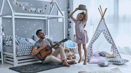 Young father plays guitar for his little daughter while she is jumping in bedroom, displaying her unique identity.