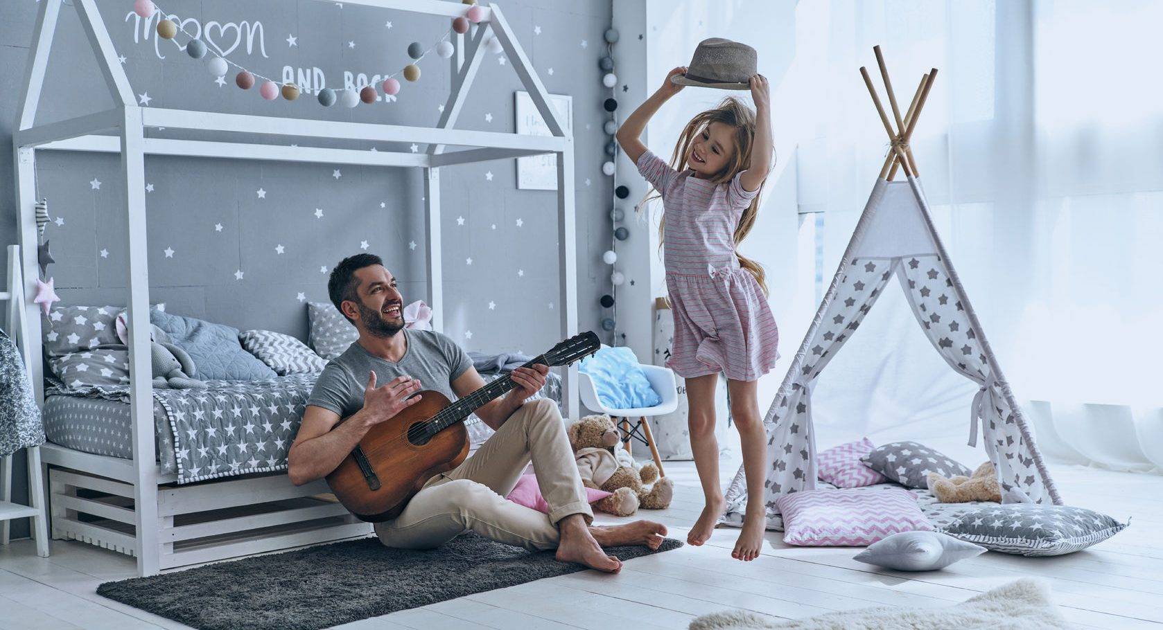 Young father plays guitar for his little daughter while she is jumping in bedroom, displaying her unique identity.
